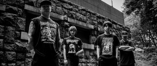 Vier Personen in dunkler Kleidung stehen vor einer Backsteinmauer. Sie blicken ernst und tragen Band-T-Shirts.