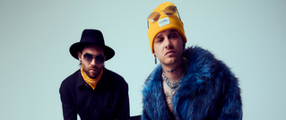 Zwei Männer in modischer Kleidung, einer trägt einen gelben Beanie und eine blaue Jacke, der andere einen schwarzen Hut und Schal.