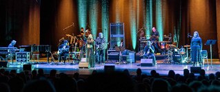 Eine Band spielt auf einer beleuchteten Bühne mit verschiedenen Musikinstrumenten. Im Vordergrund schaut ein Publikum zu.
