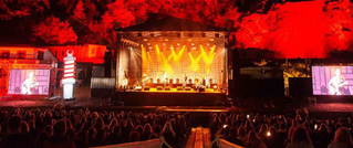 Ein Konzert bei Nacht mit orange beleuchteter Bühne, flankiert von großen Bildschirmen. Zuschauer sitzen im Vordergrund.