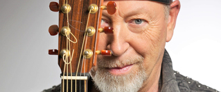 Ein Mann mit Bart und Mütze, lächelt sanft, während er eine Akustikgitarre vor sein Gesicht hält.