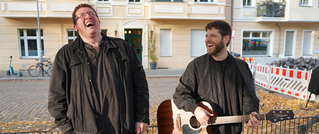 Zwei lachende Männer stehen draußen. Einer hält eine Gitarre. Im Hintergrund ist ein Gebäude und eine Baustelle zu sehen.