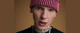 Junger Mann mit rosa Mütze, Gesichtspiercings und Hals-Tattoos vor beigem Hintergrund schaut direkt in die Kamera.