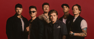 Sechs Männer stehen vor einem roten Hintergrund. Sie tragen lässige Kleidung, teils mit Tattoos und modischen Accessoires.