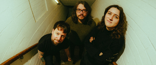 Drei Personen stehen auf einer Treppe in einem weißen, schmalen Flur. Sie blicken nach oben zur Kamera und wirken entspannt.