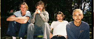 Vier Personen sitzen entspannt auf einer Bank im Freien, umgeben von Bäumen. Alle haben Schnurrbärte und schauen direkt in die Kamera.