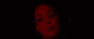 Porträt einer Frau in rotem Licht, mit dramatischem Augen-Make-up und neutralem Gesichtsausdruck, vor dunklem Hintergrund.