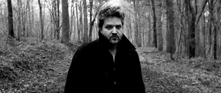 Ein Mann mit Bart und lockigem Haar steht in einem Wald. Er trägt einen dunklen Mantel. Die Umgebung ist herbstlich kahl.
