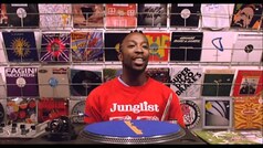Ein Mann in einem roten 'Junglist'-Shirt steht vor einer Wand voller Schallplatten. Vor ihm ist ein Plattenspieler.