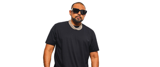 Eine Person mit Sonnenbrille und schwarzem T-Shirt steht vor einem weißen Hintergrund, trägt eine dicke silberne Kette.