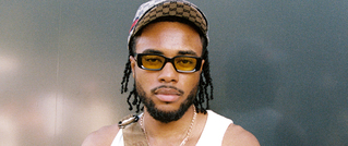 Eine Person mit Brille und Cap trägt ein weißes Tanktop und schaut in die Kamera. Hintergrund ist schlicht und dunkel.