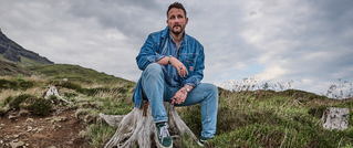 Ein Mann in Jeans und Jeansjacke sitzt auf einem Baumstumpf in einer hügeligen, grasbewachsenen Landschaft. Der Himmel ist bewölkt.