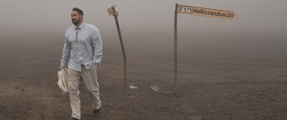 Ein Mann geht auf einem nebligen, kargen Weg an einem umgebogenen Schild vorbei, das F575 Hellsisandur zeigt.