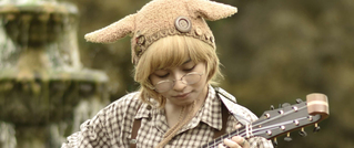 Person mit Brille und flauschiger Tiermütze spielt Gitarre im Freien. Hintergrund zeigt unscharfen, natürlichen Bereich.