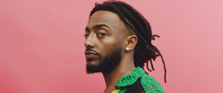 Ein Mann mit Dreadlocks und Bart steht vor einem rosa Hintergrund. Er trägt ein grünes und gelbes Oberteil und schaut ernst zur Seite.