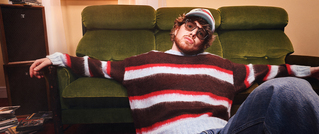 Ein Mann mit Brille und Mütze sitzt lässig mit ausgestreckten Armen auf einem grünen Sofa. Er trägt einen gestreiften Pullover.