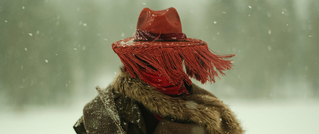 Person mit rotem Fransenhut und Fellmantel steht im Schneefall. Hintergrund unscharf und verschneit.