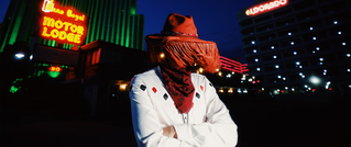 Person in weißem Hemd und rotem Cowboyhut steht nachts vor Neonlichtern eines Motels. Gesicht durch Fransen verdeckt.