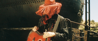 Person mit rotem Hut und Gitarre steht vor einem alten, rostigen Metallbehälter im Freien.