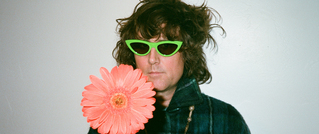 Person mit zerzaustem Haar trägt grüne Sonnenbrille, hält große, pinke Blume vor Gesicht, vor weißem Hintergrund.