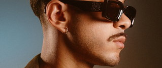 Mann im Profil mit Schnurrbart, trägt große schwarze Sonnenbrille, Ohrring und braune, kurze Haare.