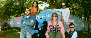 Eine Gruppe von sechs Personen posiert vor einem BBC-Banner im Freien. Eine Person trägt ein Kostüm mit blauer Katzenmaske.