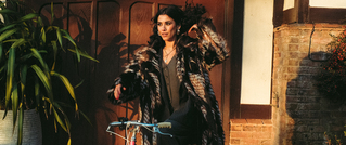 Eine Person in einem eleganten Mantel steht neben einem Fahrrad, an einem sonnigen Tag vor einer Holztür, neben einer Pflanze.