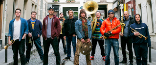 Eine Gruppe von zehn Personen steht auf einer gepflasterten Straße und hält Musikinstrumente, darunter Trompeten und eine Tuba.